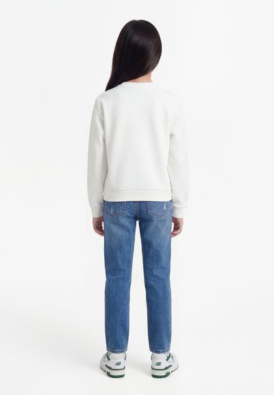 Enfant aux longs cheveux foncés portant un sweat-shirt blanc uni, un jean bleu et des baskets blanches, debout de dos sur un fond blanc.