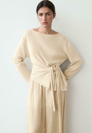 Žena nosící béžový svetr s dlouhými rukávy s páskem v pase a volné béžové kalhoty, stojící na jednobarevném světlém pozadí.