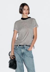 T-shirt grigia a maniche corte con colletto rotondo e finiture a contrasto, abbinata a jeans a gamba larga blu chiaro. Il modello tiene una borsa a tracolla nera con texture.