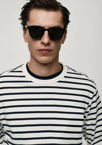 Gestreiftes Langarmshirt mit navyblauen und weißen horizontalen Streifen, das einen Rundhalsausschnitt hat. Schwarze Sonnenbrille verdeckt die Augen. Glatte Textur.