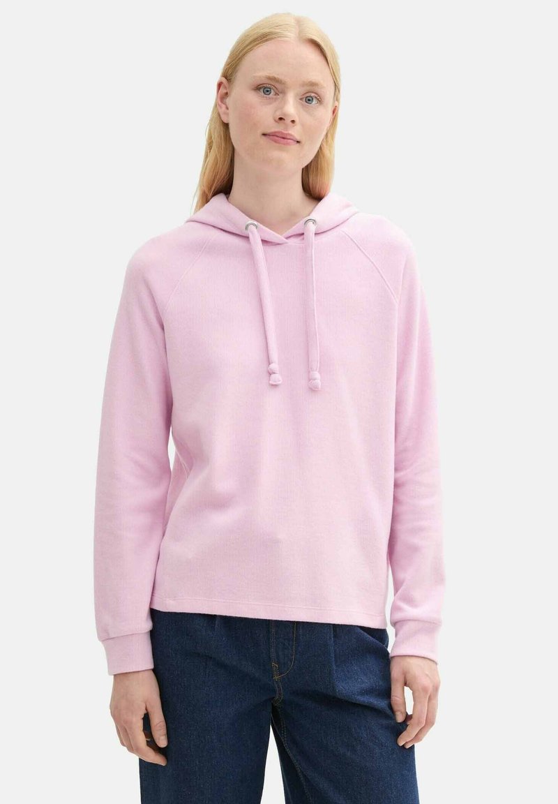 Svetloružová mikina s kapucňou na šnúrku, s rebrovanými manžetami a voľným strihom. Nosená cez tmavomodré džínsy s hladkou textúrou.