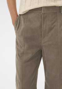 Bruine suède broek met zijzakken, gecombineerd met een lichtbeige top; een hand rust nonchalant in een zak.