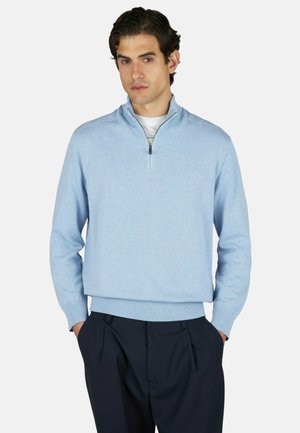 Homme portant un pull à fermeture éclair courte bleu clair par-dessus une chemise blanche avec un pantalon foncé, debout les mains dans les poches sur un fond uni.