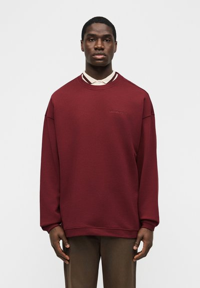 Νεαρός άνδρας που φοράει μεγαλύτερου μεγέθους μπουφάν σε μπορντό χρώμα πάνω από ένα μπεζ πουκάμισο με γιακά, στέκεται μπροστά σε απλό ανοιχτό γκρι φόντο.