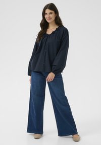 Chemisier bleu marine à boutons avec un col froncé, manches larges, associé à un jean large bleu clair et des chaussures beige.