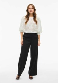 Blouse blanche texturée avec des manches bouffantes, assortie à un pantalon noir large taille haute et des talons marron. Design simple et moderne.