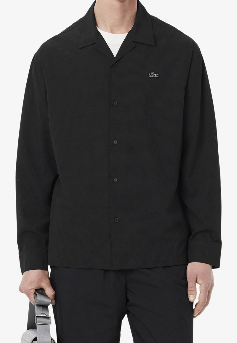 Homme portant une chemise noire à manches longues boutonnée avec un petit logo brodé sur la poitrine, tenant une sangle grise dans la main gauche.