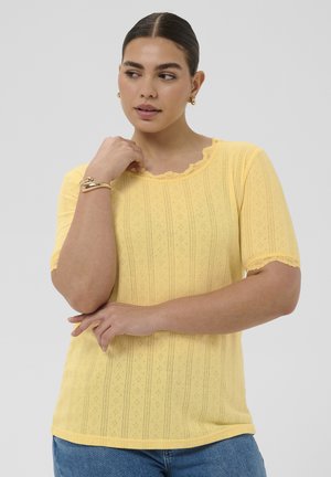 Kvinde med mørkt hår iført en gul kortærmet striktop med blondekant, iført guldbøjler og et armbånd, poserer mod en ensfarvet baggrund.