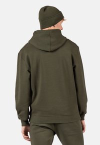 Felpa verde oliva con cappuccio ampio e polsini a costine, abbinata a un berretto a coste coordinato, design minimale, texture morbida.