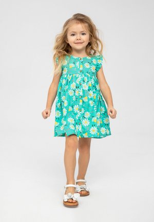 Turquoise jurk met witte madeliefjes en gele bloemafdrukken, korte rimpelrokken mouwen en een geraffineerde taille. Witte sandalen met bloemaccenten.
