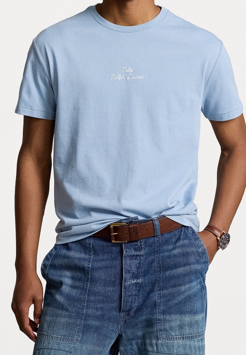 Man wearing light blue Polo Ralph Lauren t-shirt tucked into blue denim pants with brown leather belt and wristwatch on left wrist.
