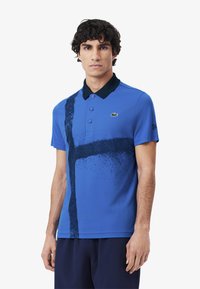 Blå polo skjorta med svart krage, med ett framträdande mörkt abstrakt mönster på framsidan. Inkluderar tre knappar och en liten logotyp.