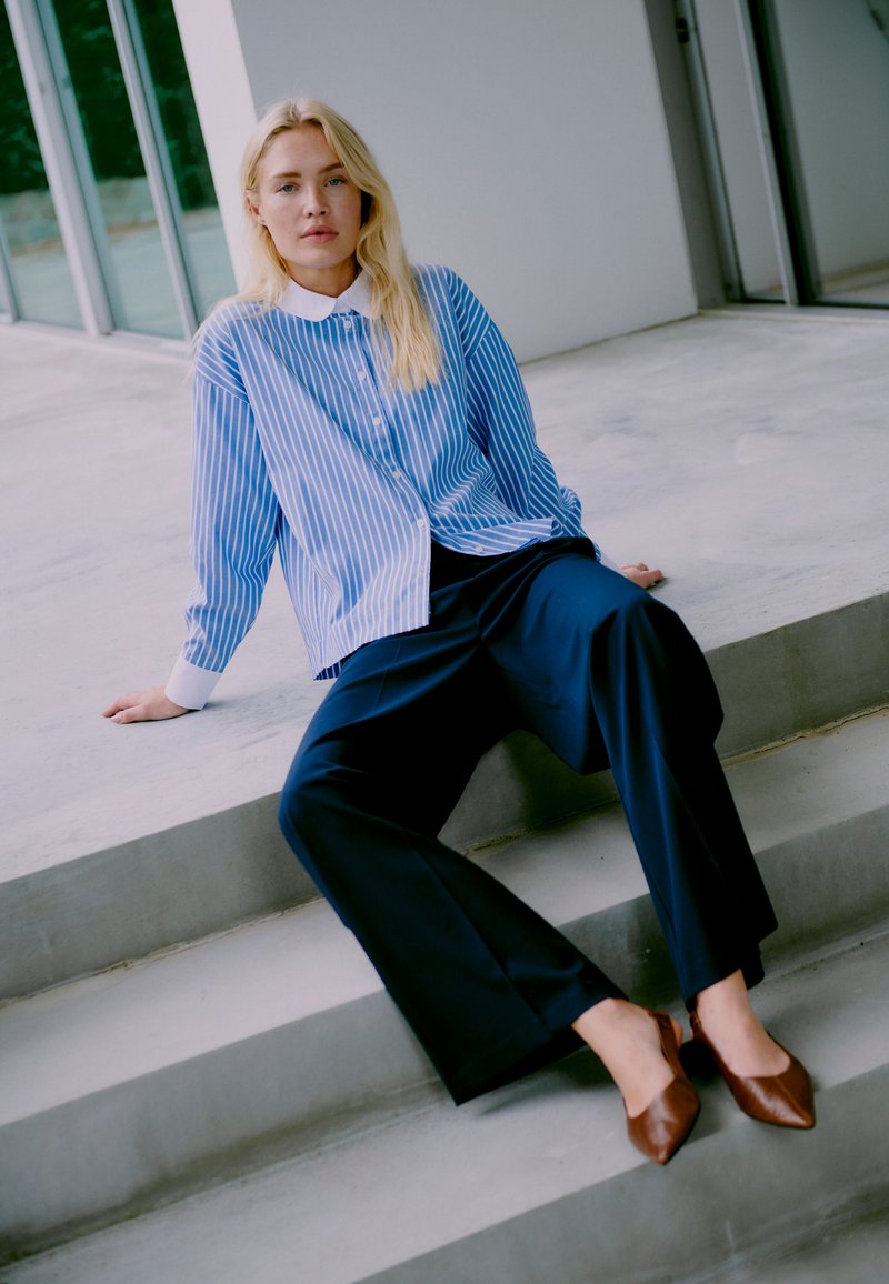Blauw en wit gestreepte blouse met een witte kraag, gecombineerd met marineblauwe wijde broek en bruine puntige flats. Betonnen trappen als achtergrond.