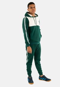 Chándal atlético verde con un panel blanco en el pecho, sudadera con cremallera, rayas laterales y acentos de logo. Combinado con zapatillas navys con rayas azules.