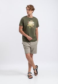 T-shirt in cotone verde oliva con una stampa grafica colorata, abbinata a pantaloni corti beige chiaro e sandali neri con accenti grigi.