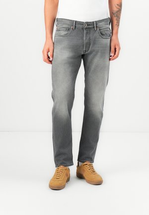 Man draagt vervaagde grijze jeans met rechte pijpen en beige veterschoenen, staat tegen een effen witte achtergrond.