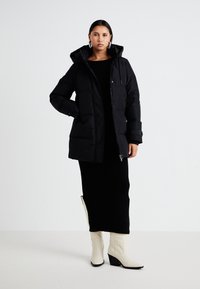 Doudoune noire avec capuche, présentant une fermeture éclair à l'avant, des poches latérales et un design matelassé. Portée sur une longue robe noire côtelée et des bottes légères.