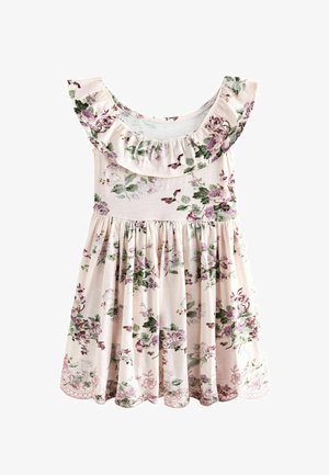 Junges Mädchen steht da, trägt ein hellrosa Blumenkleid mit Rüschenärmeln und weiße Sandalen, vor einem einfarbigen beigen Hintergrund.