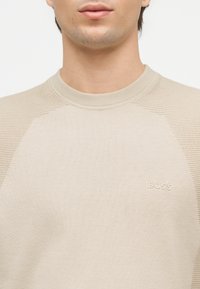 Άνδρας που φοράει μπεζ πλεκτό πουλόβερ με ριμπ ώμους και λογότυπο "BOSS" στο αριστερό μέρος του στήθους, απεικονίζεται από το λαιμό έως τον άνω κορμό.