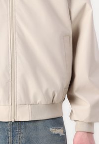Lys beige bomberjakke med lynlås foran, ribbede manchetter og kant, med en glat, let blank overflade og en sidelomme.