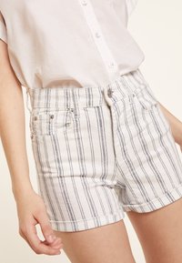 Pantalones cortos de denim de rayas blancas y azul claro con una cintura alta, que cuentan con dos bolsillos frontales y un dobladillo enrollado. El material parece ligero y texturizado.