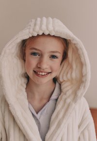 Peignoir blanc en peluche avec une capuche texturée, présentant un motif côtelé. Le peignoir a une apparence douce et confortable et est porté par-dessus une chemise claire.