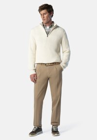 Maglione bianco a coste con zip a metà, indossato sopra una camicia a quadretti grigia. I pantaloni beige e le sneaker scure con lacci a contrasto completano il look.