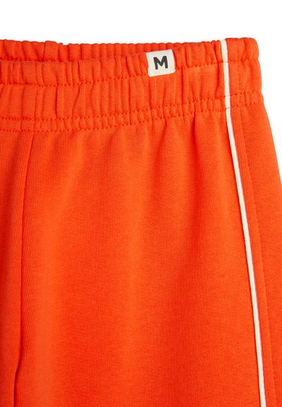 Oransje joggebukser med elastisk midjebånd, hvit sidestripe og størrelseetikett "M" sydd på midjebåndet. Myk tekstur.