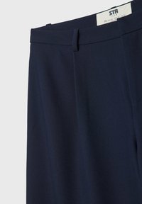 Pantalon sur mesure bleu marine avec passants de ceinture et une étiquette de marque visible à l'intérieur de la ceinture indiquant la taille et les détails d'origine.