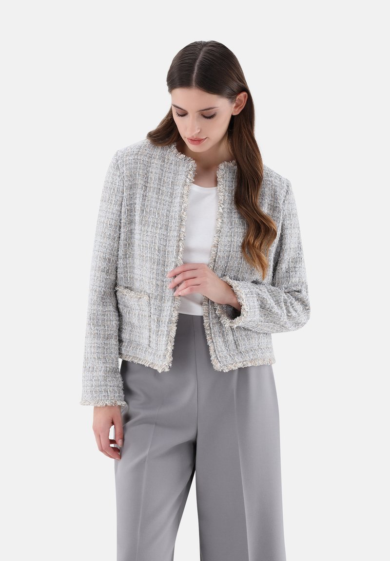 Hellgraue Tweedjacke mit fransigen Kanten, rundem Ausschnitt und zwei Fronttaschen, kombiniert mit einem weißen Oberteil und grauen Hosen.