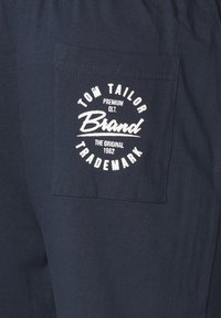 Pantalon en coton bleu marine avec une poche patch arborant un logo imprimé en blanc : "TOM TAILOR PREMIUM QLT. The Original 1962." Détails de couture simples.