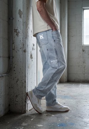Personne portant un jean en denim bleu clair avec le texte blanc "K" et "KANI", baskets beiges, debout et appuyée contre un mur en béton usé dans une pièce vide.