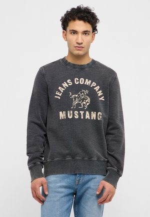 Junger Mann mit lockigem Haar, der ein verblasstes schwarzes Sweatshirt mit der Aufschrift "Jeans Company Mustang" und einem Rodeo-Reiter-Logo trägt, kombiniert mit hellblauen Jeans.