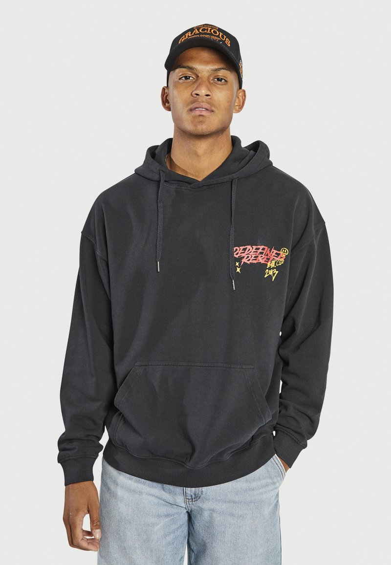 Schwarzer Hoodie mit Fronttasche, Kordelzugkapuze und grafischem Text in Pink und Gelb auf der linken Brust. Getragen mit hellblauen Jeans.