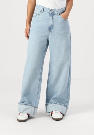 Jeans larges en bleu clair avec une taille haute, des ourlets retroussés et des poches latérales, fabriqués en tissu denim. Portés avec des baskets blanches.