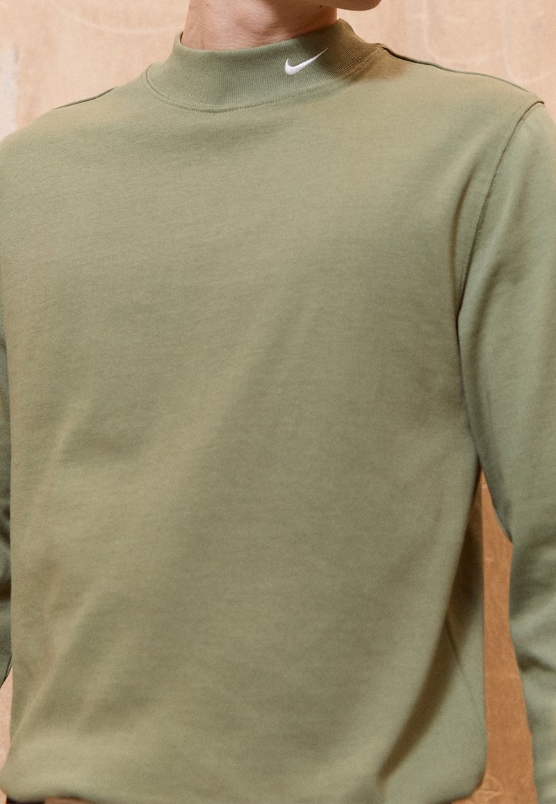 Gros plan sur une personne portant un sweat-shirt Nike vert olive avec un logo Nike blanc sur le col.