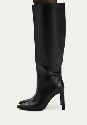 Stivali neri in pelle al ginocchio con punte appuntite e tacchi a spillo alti indossati da una modella su sfondo neutro.