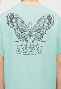 Πίσω μέρος ενός γαλάζιου πουκαμίσου με ένα μεγάλο, μαύρο, λεπτομερές γραφικό πεταλούδας πάνω από τη λέξη "Jackher" και μικρότερο κείμενο από κάτω.