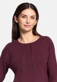 Bordowy sweter z okrągłym dekoltem, z dwoma plisami z przodu i gładką fakturą. Długie rękawy i luźny krój.