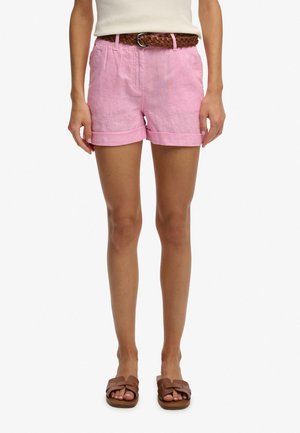 Mujer con pantalones cortos rosas con dobladillo enrollado y cinturón trenzado marrón, camiseta blanca sin mangas y sandalias marrones tipo "slide", de pie recta.