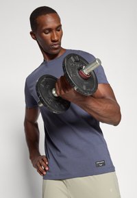 Grå treningstrøye med korte ermer, som holder en svart vektplate hantel. Lysegrå shorts synlige, med en liten logo på siden.