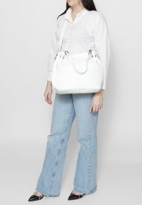 Sac à main en cuir blanc avec double poignée et bandoulière ajustable, présentant une forme arrondie et des accents métalliques. Surface texturée, design décontracté.