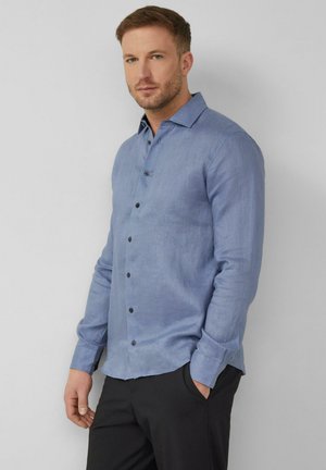 Mężczyzna z krótkimi włosami i lekkim zarostem, ubrany w jasnoniebieską koszulę z długim rękawem i czarne spodnie, stojący na tle gładkiego tła.