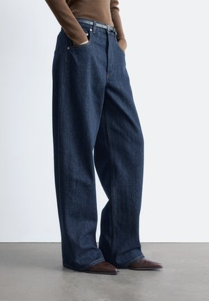 Person wearing loose dark blue jeans, brown belt, brown long-sleeve top, and pointed brown shoes standing on a gray floor.