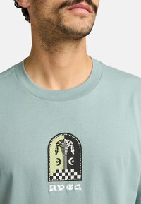 Camiseta de cuello redondo en verde menta con un gráfico en blanco y negro de un jarrón con plantas, patrón de tablero de ajedrez y el texto "RYEGA" en blanco.