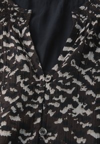 Blouse à motifs marron et noir avec un tissu plissé, des épaules smockées et deux boutons visibles sur le devant.