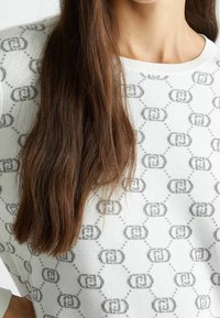 Maglione bianco con un motivo logo grigio ripetuto, colletto a costine e texture morbida. Capelli che coprono parzialmente il capo.
