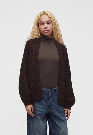 Mladá žena s dlhými kučeravými blond vlasmi, oblečená v hnedom roláku a tmavohnedom pletenom kardigáne s modrými džínsami na jednoduchom pozadí.