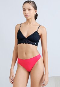 Bralette de encaje negro con bordes festoneados; combinado con bragas rojas de bordes festoneados. Ambas prendas presentan una textura suave y un diseño minimalista.