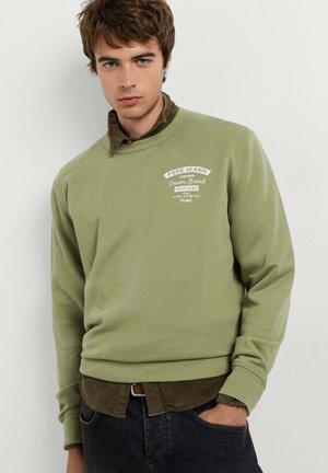Junger Mann trägt ein hellgrünes Pepe Jeans Sweatshirt über einem braunen Hemd, die Hand in der Tasche der dunklen Jeans vor einem schlichten weißen Hintergrund.